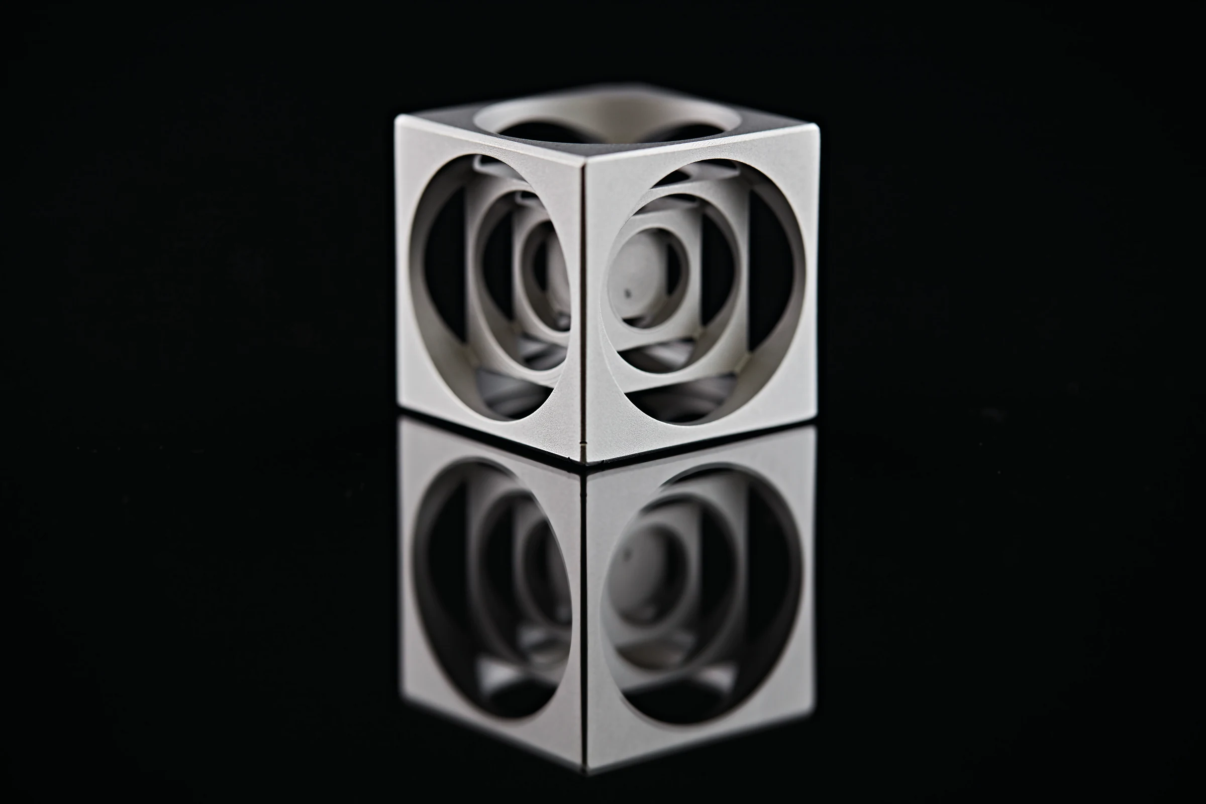 Metallischer Bauteilwürfel mit runden Öffnungen und gleichmäßig matter Oberfläche vor dunklem Hintergrund